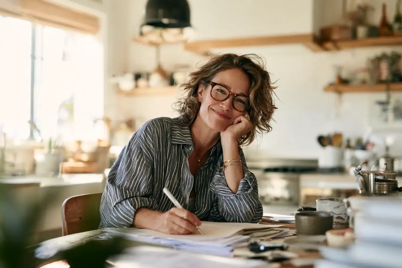 A woman in her fifties smiling while writing notes at a kitchen table before an appointment