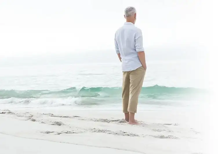 An older man standing barefoot on a beach, looking out at the waves