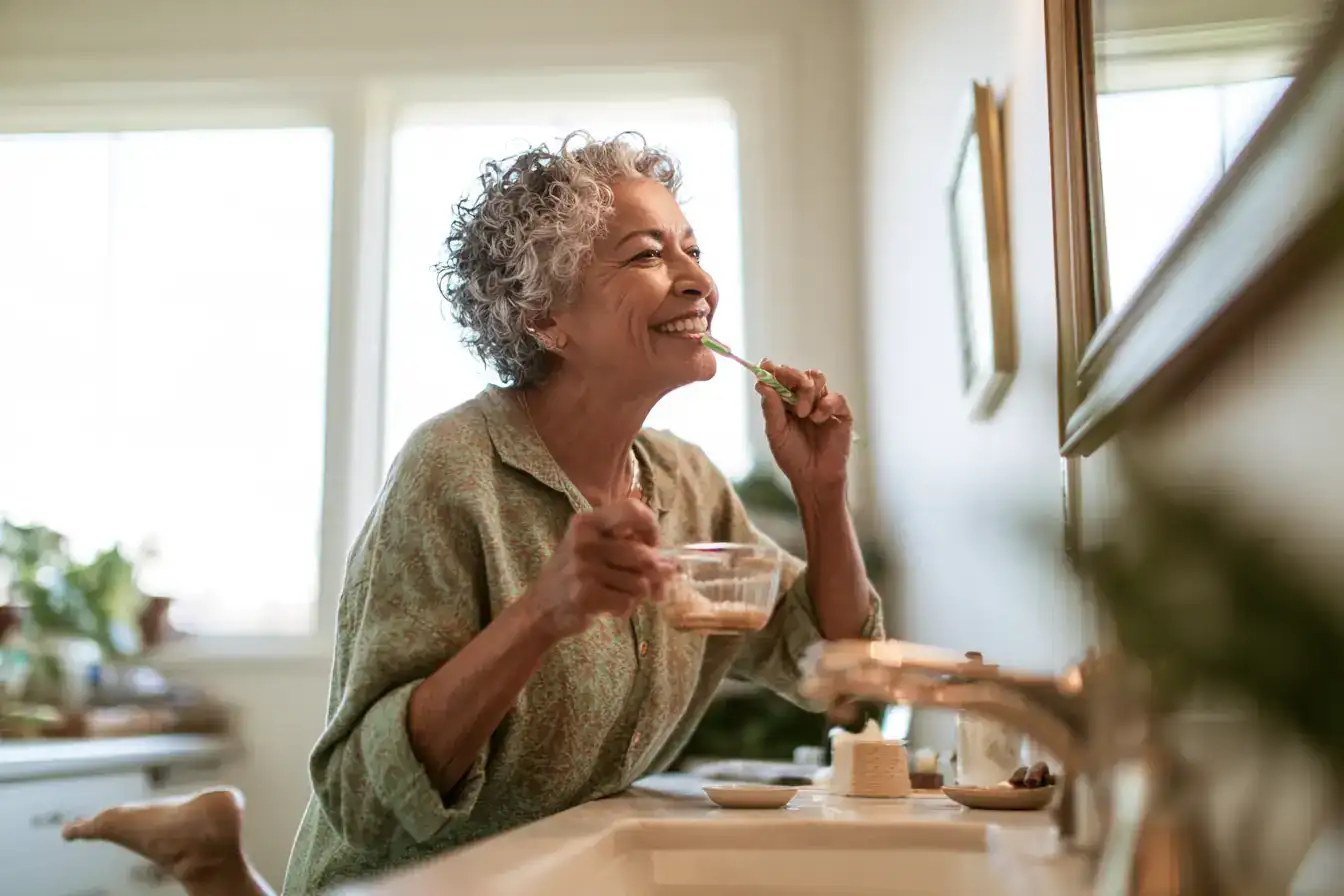 A Latina woman in her seventies standing on one leg at the bathroom sink, brushing teeth